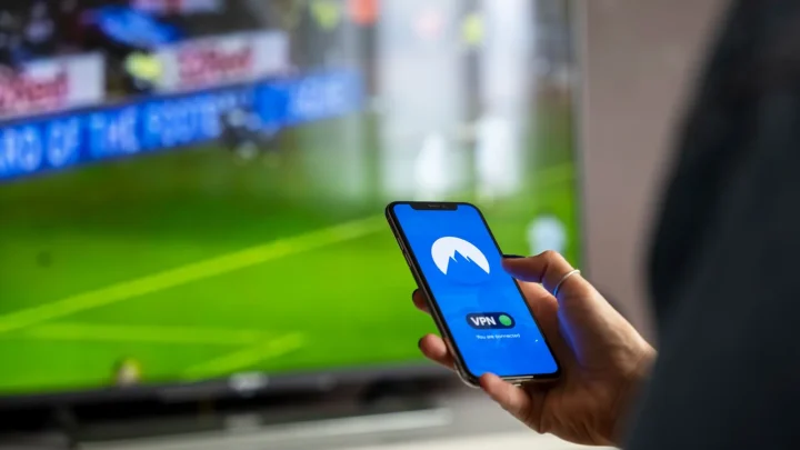Close-up of a person holding a smartphone with a VPN app, streaming sports on TV.