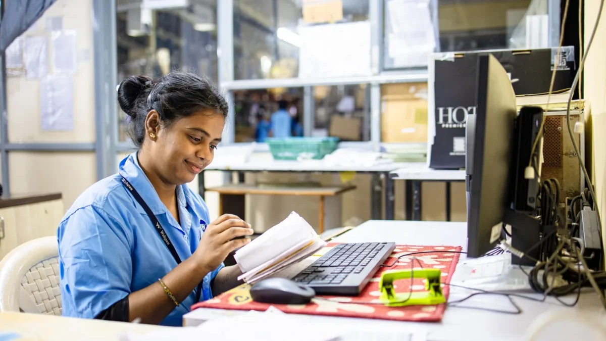 Smiling woman in blue uniform working at office desk with computer and paperwork.