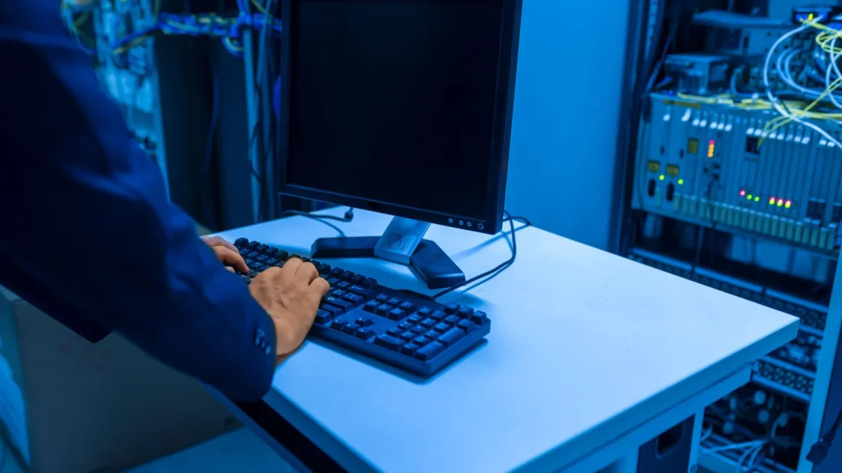 An IT professional operates a computer in a server room, managing network systems and connected devices.